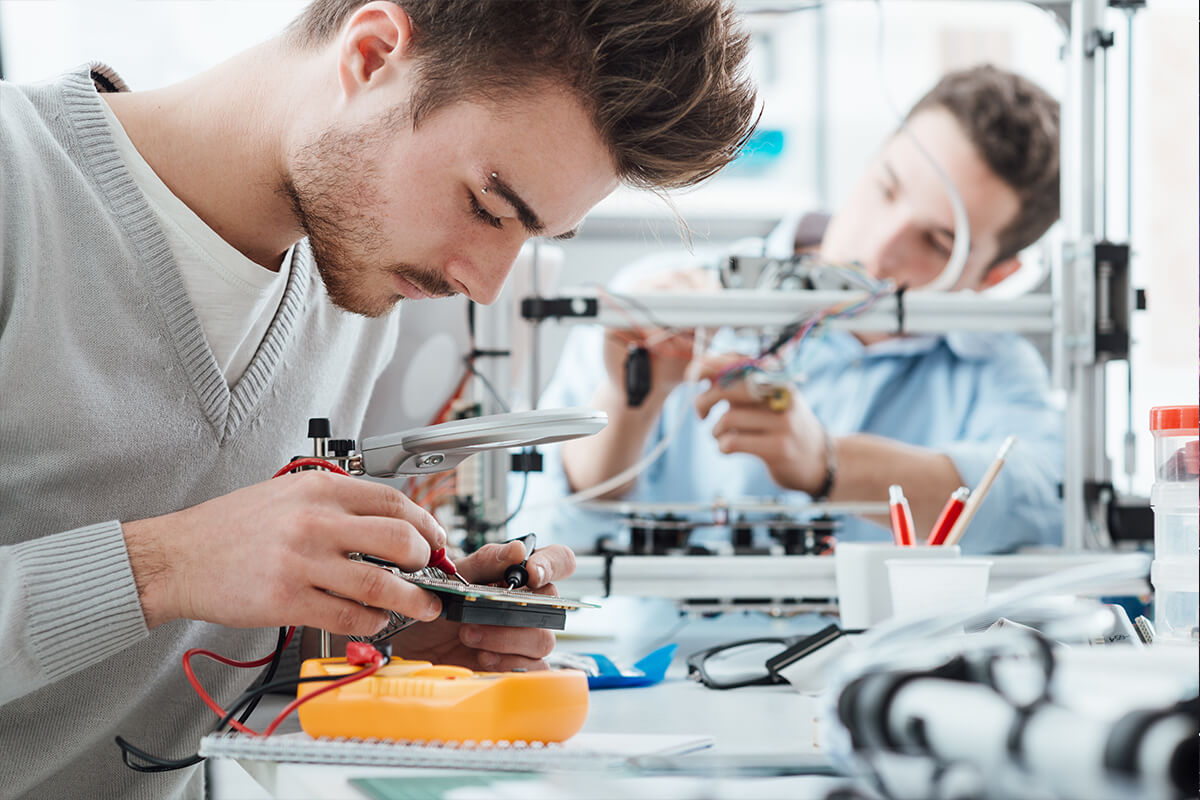 Two male engineers working in a lab with voltage and current tester and a 3D printer